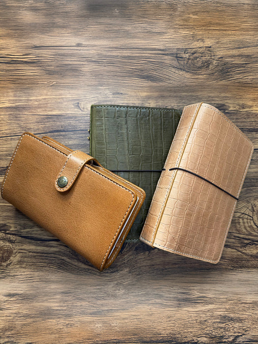 Alt Text:
Three handcrafted leather journals displayed on a wooden background, showcasing different closure styles: a tan journal with a snap closure, a green crocodile-textured journal with no visible closure, and a beige crocodile-textured journal with an elastic band.