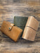 Alt Text:
Three handcrafted leather journals displayed on a wooden background, showcasing different closure styles: a tan journal with a snap closure, a green crocodile-textured journal with no visible closure, and a beige crocodile-textured journal with an elastic band.